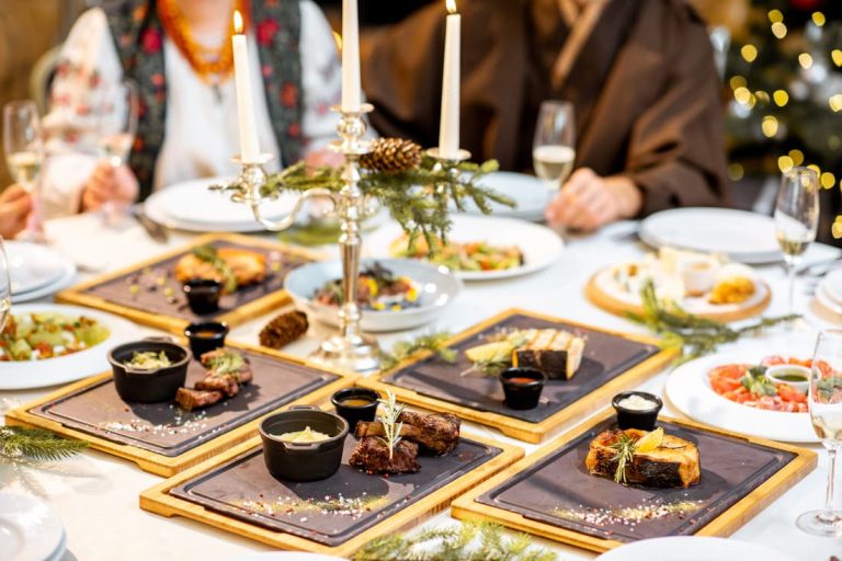 L'image montre une table de dîner festif pour le Nouvel An, avec des bougies, des plats gastronomiques sur des sets en ardoise et un sapin de Noël en arrière-plan, créant une ambiance chaleureuse et conviviale.